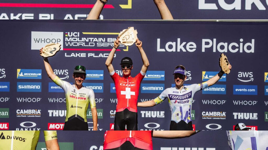 Finn Treudler jubelt auf dem Podium und hält stolz die Trophäe für seinen U23-Triumph in Lake Placid in die Höhe. Finn Treudler celebrates on the podium, proudly raising the trophy for his U23 triumph in Lake Placid.