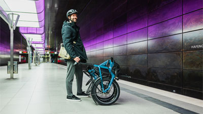 A man is standing at a subway station. In front of him is a folded blue folding bike.