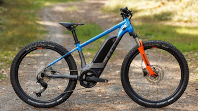 The image shows a gray Cube children's e-bike with blue and orange details. It is standing on a forest path.
